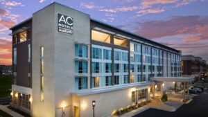 Modern exterior of the AC Hotels by Marriott building at dusk, with a stunning purple and orange sunset sky, illuminated entrance, and lush greenery.
