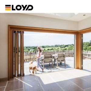 A girl and a golden retriever running through a fully opened ELOYD wooden-framed folding glass door from a bright interior to a stone patio with outdoor furniture, overlooking a green field.
