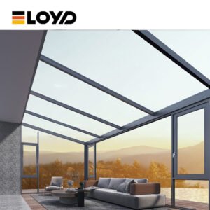 Modern ELOYD sunroom with a dark gray frame, full-height transparent glass walls and ceiling, a light gray sofa with brown cushions, and a view of mountains and trees.
