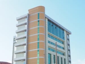 Modern multi-story office or commercial building with a blue glass curtain wall, beige and yellow tile sections, and silver horizontal accents under a clear blue sky.
