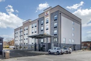 Modern exterior of the Comfort Suites hotel building, a multi-story structure in light gray and beige with dark stone accents, a covered entrance, and a parking lot with cars under a blue sky.