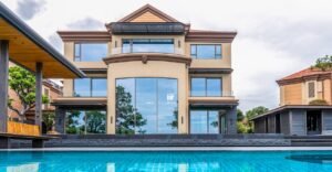 Luxurious modern three-story villa with beige walls, large windows, a brown roof, and a prominent blue swimming pool in the foreground.