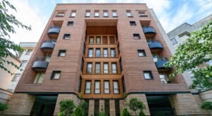A modern seven-story red brick apartment building with a recessed central facade, black metal-framed balconies, some with arched canopies, and a light-colored stone base.