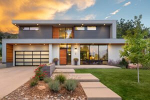 A modern two-story residence with white walls, dark grey metal finishes and wooden louvers. Large floor-to-ceiling windows, black checkered garage door, a stone path leading to the front door, a garden with flowers, and a backdrop of a wooded area at dusk.