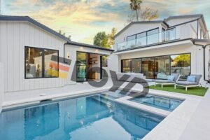 A modern-style white single-family house with large glass windows. In front of the house is a blue swimming pool, gray lounge chairs and green artificial lawn. The atmosphere is peaceful at dusk.