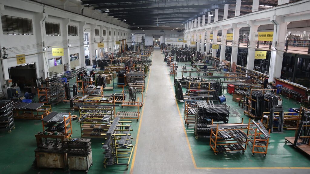 A clean and organized sunroom production area inside a factory, showing orange and black material racks, workbenches with metal components, and yellow safety markings on the green floor.