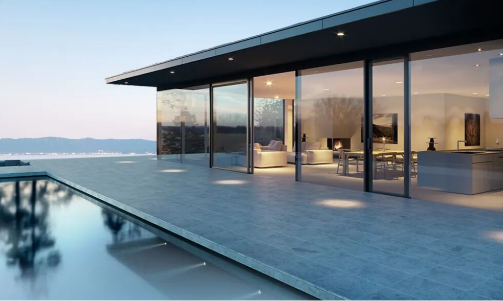 A contemporary house exterior featuring large floor-to-ceiling glass windows that reveal an interior with sofas, tables, and a fireplace, set against a rectangular swimming pool with clear blue water, and backdrop of hazy mountains and sky, emphasizing minimalist and serene architecture.