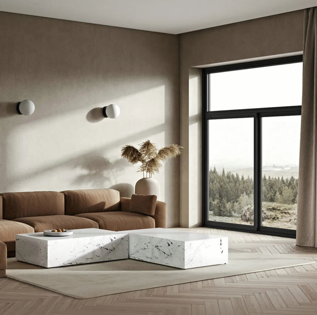 A cozy modern minimalist living room featuring a brown fabric sofa, two white marble coffee tables with a fruit bowl, light brown walls with circular sconces on each side, a beige vase with dried grass near the window, and a large black-framed window offering serene views of a dense forest. The space includes light wood flooring and a carpet, creating a warm, tranquil, and comfortable atmosphere.